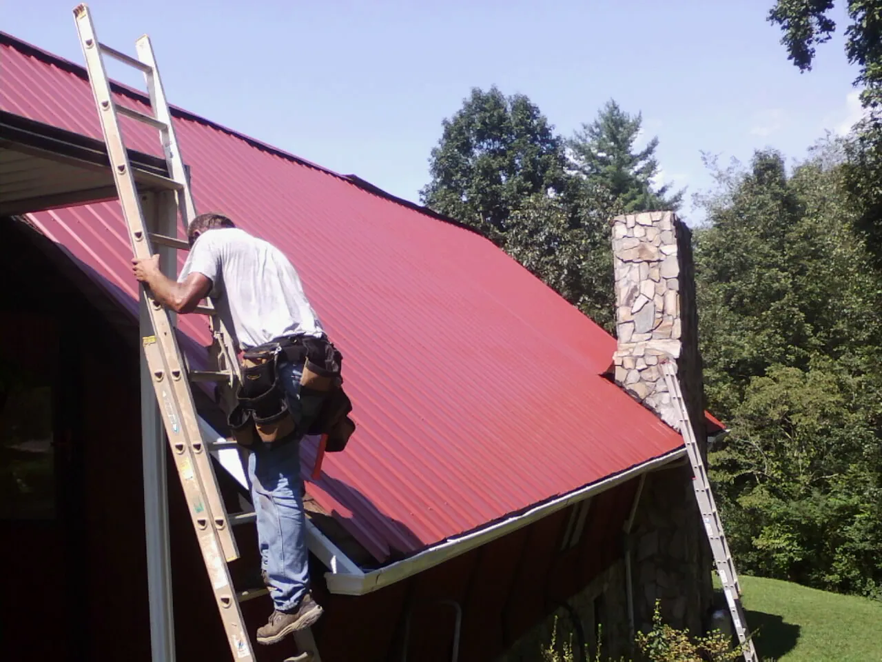 Roofer on a ladder beside a red metal roof and stone chimney — Soffit & Fascia Repair by Roofing Brothers in Newborn